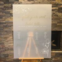 A frosted acrylic seating chart for weddings which is semi transparent. It features white lettering with gold headings and floral detail