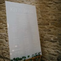 A frosted acrylic seating chart for weddings which is semi transparent. Placed on a wooden easel. It features white lettering.